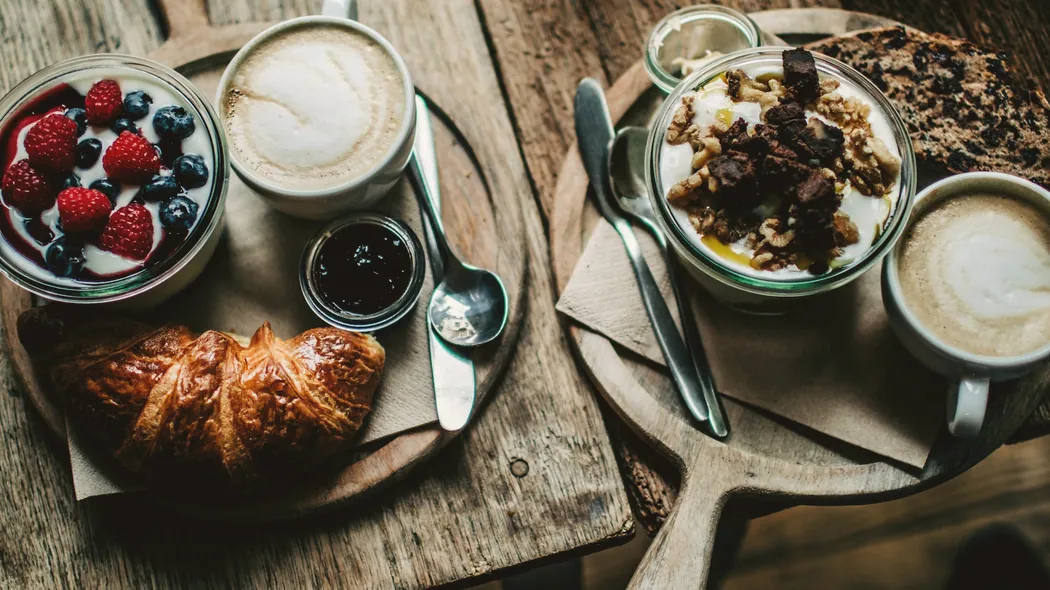 Frühstückstisch mit Croissant, Beeren, Joghurt und zwei Cappuccino auf dunklem Holz.
