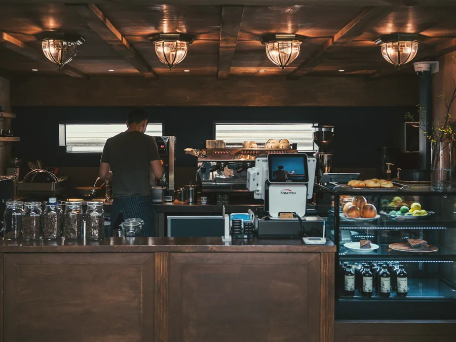 Blick auf den Tresen des Cafés mit Auslage, Kaffeemaschine und einer Person bei der Arbeit.