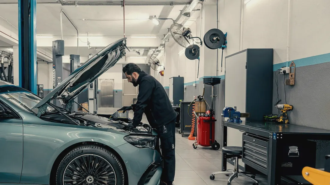 Mechaniker arbeitet in einer hellen Werkstatt an der geöffneten Motorhaube eines Fahrzeugs.
