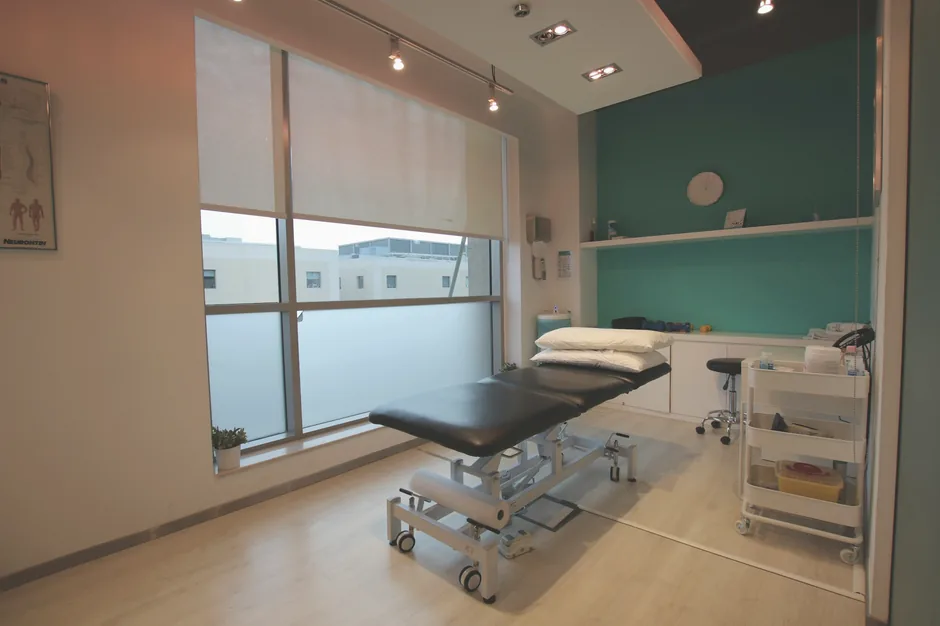 Heller Behandlungsraum mit Therapieliege, Fenster und Materialwagen.